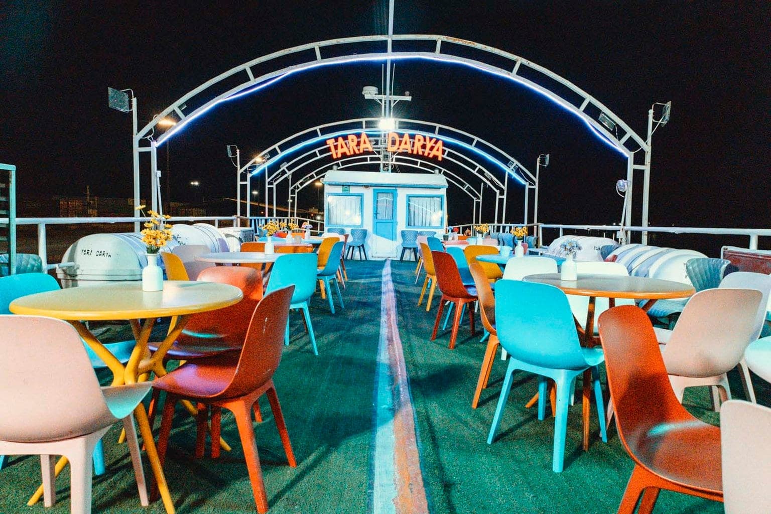 عرشهٔ باز کشتی تارا قشم در شب با قوس‌های فلزی نوری، تابلوی نئون TARA DARYA و میز و صندلی‌های رنگی خالی