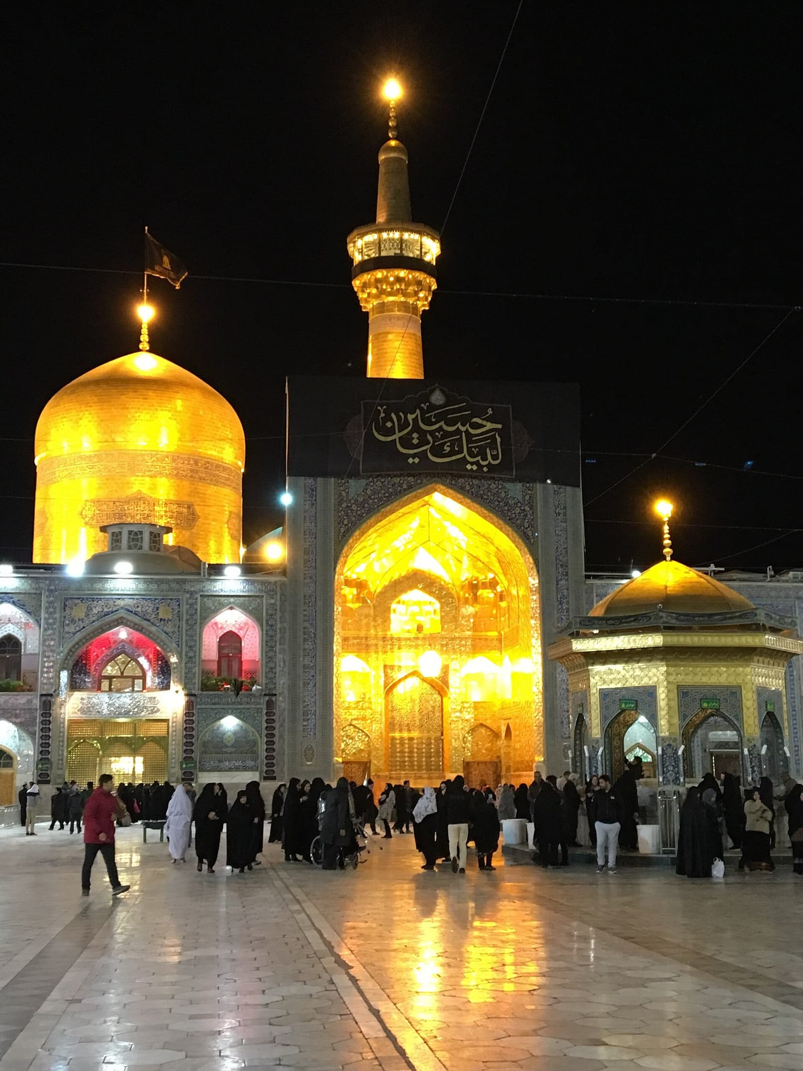 نمای شبانه صحن حرم رضوی با گنبد طلایی، گلدسته زرین و ایوان درخشان و رفت‌وآمد زائران