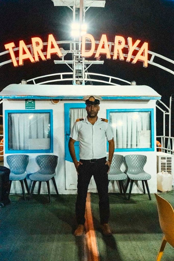 ناخدای کشتی تارا دریا قشم ایستاده مقابل کابین سفید و آبی با قوس نورانی TARA DARYA در شب