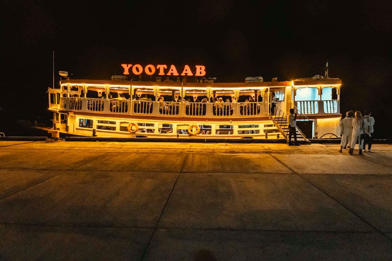 نمای دور کشتی یوتاب قشم از اسکلهٔ تاریک در شب — کل بدنه با ریسه و حروف YOOTAAB روشن شده