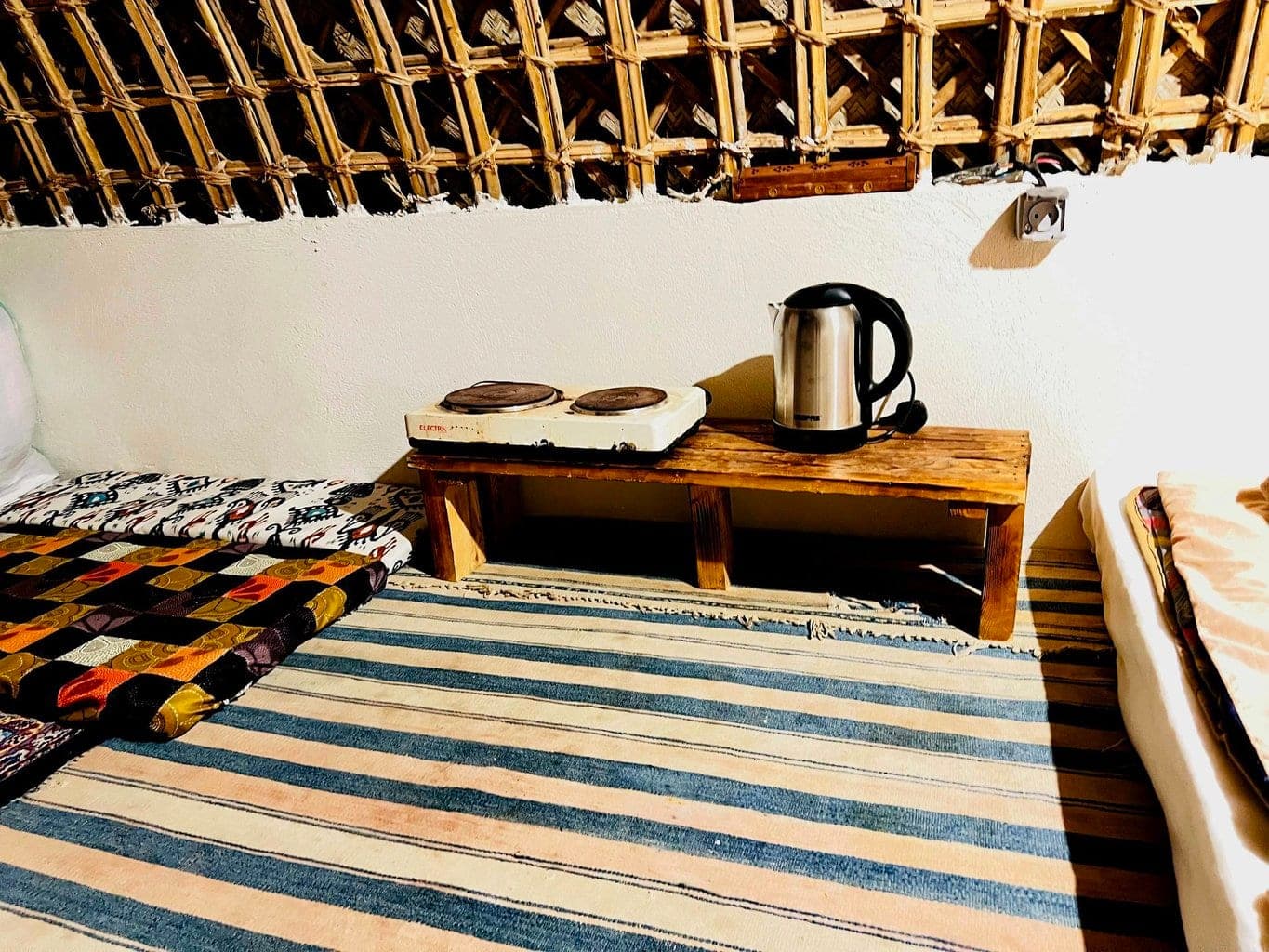 گوشهٔ دم‌نوش با اجاق برقی و کتری روی نیمکت چوبی و کف‌پوشِ گلیم در بوم‌گردی کاژه هنگام